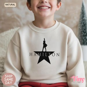 Peut inclure: Un sweat-shirt beige avec une silhouette noire d'une personne sur une étoile et le mot "HAMILTON" en dessous. Le sweat-shirt est de couleur neutre et a un aspect doux. Le texte "NATURAL" et "EXPÉDITION LE JOUR MÊME" est également inclus.