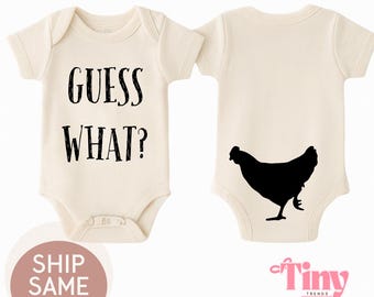 May include: A cream-colored baby onesie with the text "GUESS WHAT?" on the front and a black silhouette of a chicken on the back.