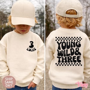 Puede incluir: Sudadera color crema con el texto "3 Lucas" en la parte delantera y "Young Wild & Three" en la trasera. El diseño incluye un patrón de cuadros negros y se combina con una gorra de béisbol a juego. Un niño lleva el conjunto.