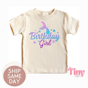 May include: Cream-colored t-shirt featuring a mermaid tail and the words "Birthday Girl" in blue and purple glitter. The design includes stars and is perfect for a birthday celebration.
