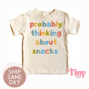 May include: Cream-colored t-shirt with the phrase "probably thinking about snacks" in colorful letters. The shirt is a short-sleeved crew neck style, perfect for kids. The text is in a playful, multi-colored font.