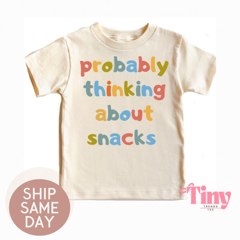 May include: Cream-colored t-shirt with the phrase "probably thinking about snacks" in colorful letters. The shirt is a short-sleeved crew neck style, perfect for kids. The text is in a playful, multi-colored font.
