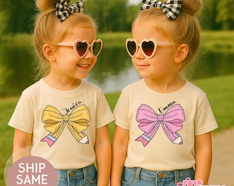 Könnte beinhalten: Zwei junge Kinder tragen herzförmige Sonnenbrillen und passende cremefarbene T-Shirts mit Schleifen- und Bleistiftmotiven. Auf einem Shirt steht "Jessica" und auf dem anderen "Emma". Beide Kinder haben ihre Haare zu Duttfrisuren mit schwarz-weiß karierten Schleifen hochgesteckt.