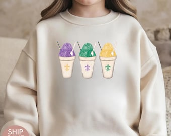 Puede incluir: Sudadera color crema con un gráfico de tres granizados coloridos: morado, verde y amarillo. Cada granizado está en un vaso blanco con un diseño de flor de lis y una pajita a rayas. La palabra "NATURAL" está impresa en la parte superior izquierda.