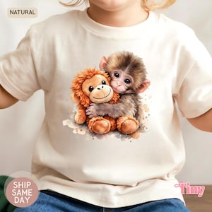 Puede incluir: Una camiseta color crema con una ilustración de acuarela de dos monos abrazándose. Un mono es marrón y el otro es naranja. La palabra "NATURAL" está impresa en la parte superior izquierda de la camiseta. Las palabras "SHIP SAME DAY" están impresas en una pegatina circular.