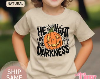 May include: A beige t-shirt featuring a smiling orange pumpkin with a cross and the text "HE IS THE LIGHT IN THE DARKNESS JOHN 1:5". The shirt has a round neck and short sleeves. The word "NATURAL" is printed on the top left.