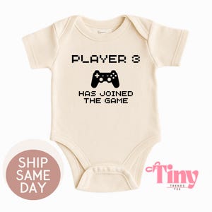 May include: Cream baby bodysuit featuring the text "PLAYER 3 HAS JOINED THE GAME" and a black controller graphic. A playful design for a newborn, ideal for a family of gamers.