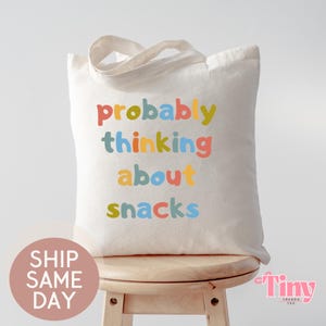 May include: A cream-colored tote bag with the phrase "probably thinking about snacks" printed in colorful letters. The bag has a simple design with a handle and is sitting on a wooden stool.