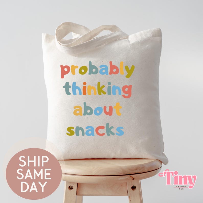 May include: A cream-colored tote bag with the phrase "probably thinking about snacks" printed in colorful letters. The bag has a simple design with a handle and is sitting on a wooden stool.