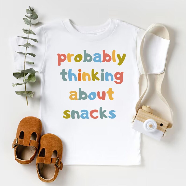 May include: White t-shirt with the phrase "probably thinking about snacks" in colorful letters. The image includes a pair of brown leather baby shoes and a small wooden toy camera.