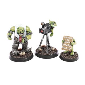 May include: Three miniature fantasy figures. One is a green-skinned orc in a white shirt and tie, holding a microphone. Another is a goblin operating a black camera on a tripod. The third figure is a goblin holding a sign that reads "REMEMBER, LIVE, LAUGH, WAAAAAGH YOU GITS!"