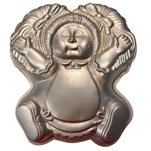 May include: A silver-colored metal cake pan in the shape of a doll with pigtails and a bow. The doll has a round face, large eyes, and a small smile. The pan is designed to create a cake in the shape of a doll.
