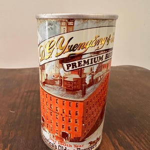 May include: Vintage D.G. Yuengling & Son Premium Beer can. The can features a detailed illustration of a brewery with red brick walls and a white banner with the brand name. The text "America's Oldest Brewer" is also visible.