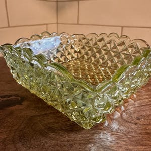 May include: A square, yellow-green glass bowl with a scalloped rim. The bowl has a diamond pattern on the sides and bottom. The bowl is sitting on a wooden surface.