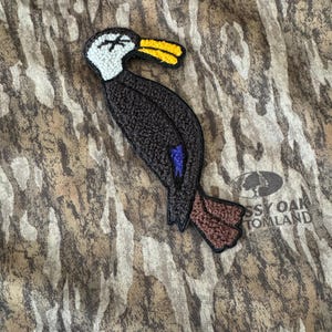 May include: A black and brown embroidered patch of a duck with a yellow beak and blue accents. The patch is on a camouflage fabric background with the text "Mossyoak Tomland".