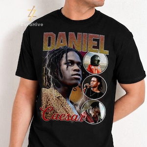 May include: A black t-shirt featuring a large graphic print of Daniel Caesar. The design includes a prominent portrait, the name DANIEL in bold letters, the name Caesar in red script, and three circular insets showing various images of the artist.