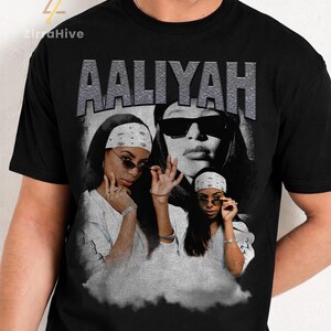 May include: Black t-shirt with a grayscale image of Aaliyah, and the name "AALIYAH" in a textured, silver font. The image shows Aaliyah wearing sunglasses and a bandana. The shirt has a classic crew neck design.