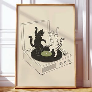 Könnte beinhalten: Ein gerahmter Kunstdruck mit einer Strichzeichnung eines Vintage-Plattenspielers auf cremefarbenem Hintergrund. Zwei Katzen, eine schwarze und eine helle mit Streifen, stehen auf der sich drehenden schwarzen Vinylplatte und tanzen.