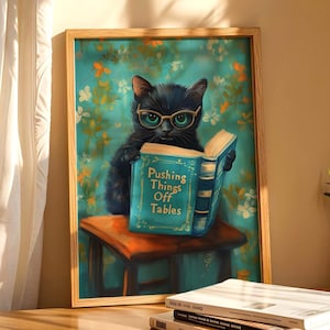 Könnte beinhalten: Ein gerahmter Wandkunstdruck zeigt eine schwarze Katze mit goldener Brille, die auf einem Holzhocker sitzt und ein offenes blaues Buch hält. Auf dem Buchdeckel steht der Text Pushing Things Off Tables in goldener Schrift vor Blumen.