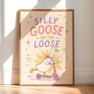 Op de afbeelding: Een ingelijste kunstprint met een witte gans die een roze sjaaltje met stippen draagt en op een roze step rijdt. De tekst luidt SILLY GOOSE ON THE LOOSE in paarse en roze letters. De achtergrond is crème met gele sterren en fonkelingen.