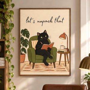 Puede incluir: Una lámina de arte mural enmarcada que muestra a un gato negro sentado en un sillón verde leyendo un libro. El texto "let's unpack that" está escrito sobre el gato. Junto al sillón hay una pequeña mesa de madera con una lámpara y una taza.