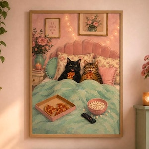 May include: A framed wall art print depicting a black cat and a tabby cat sitting in a bed with a teal blanket, eating pizza slices. A pizza box with remaining slices, a bowl of popcorn, and a remote control rest on the bed. The wall features pink decor.