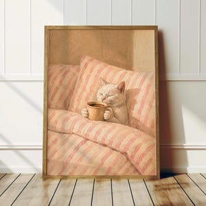 May include: A framed wall art print depicting a white cat with closed eyes, nestled under a pink and white striped blanket. The cat holds a tan ceramic mug with both paws. The scene is rendered in a soft, warm, painterly style with light tones.