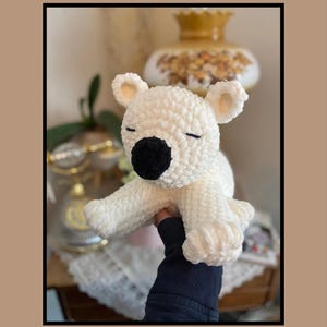 Könnte beinhalten: Weißes gehäkeltes Benson Bear Häkelmuster. Der Bär hat schwarze Augen und eine schwarze Nase. Der Bär sitzt auf einer Hand.