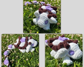 May include: A crochet pattern for a baby dragon named Clover. The dragon is white with brown and purple accents. The image includes three different views of the finished dragon, set against a background of green grass and white flowers. The text "Clover the Baby Dragon" and "Crochet Pattern" are also included.
