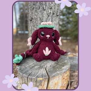 May include: Whimsy Crochet Pattern: A crocheted amigurumi toy with a burgundy body, green and pink flower accents, and a pink and green hat. The toy is sitting on a tree stump.