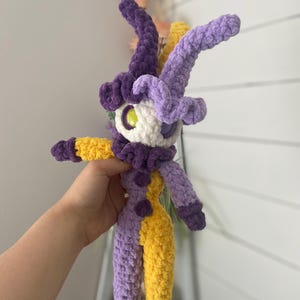 Pu&ograve; includere: Una bambola all'uncinetto fatta a mano con un design stravagante. La bambola presenta una combinazione di colori viola e giallo, con un viso bianco e grandi occhi. Ha lunghe corna curve e un colletto a balze. La bambola &egrave; alta circa 25 cm.