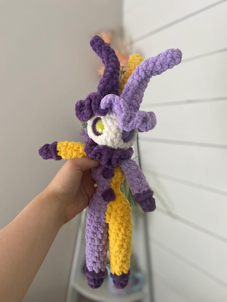 Pu&ograve; includere: Una bambola all'uncinetto fatta a mano con un design stravagante. La bambola presenta una combinazione di colori viola e giallo, con un viso bianco e grandi occhi. Ha lunghe corna curve e un colletto a balze. La bambola &egrave; alta circa 25 cm.