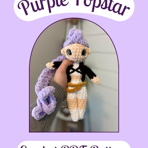 May include: A handmade crochet doll with a purple hairstyle and long braid, wearing a black top and white trousers with tan accents. The doll has large, expressive eyes. The image includes the text "Purple Popstar" and "Crochet PDF Pattern".