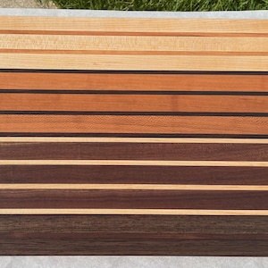 May include: A rectangular wooden cutting board with a striped pattern. The stripes alternate between light and dark brown wood tones, with some black accents. The board is likely used for food preparation.