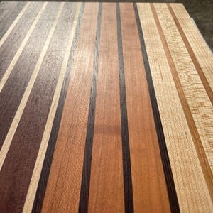 May include: Close-up of a wooden cutting board with alternating stripes of various wood tones. The board features dark brown, light brown, and natural wood colors, creating a visually appealing pattern. The wood grain is visible.