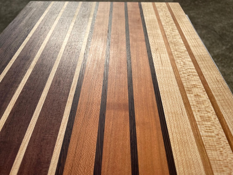 May include: Close-up of a wooden cutting board with alternating stripes of various wood tones. The board features dark brown, light brown, and natural wood colors, creating a visually appealing pattern. The wood grain is visible.