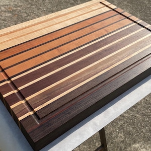 May include: A rectangular wooden cutting board with a striped design. The board features alternating stripes of light and dark wood tones, including shades of brown, tan, and black. A recessed groove runs along the edge of the board.