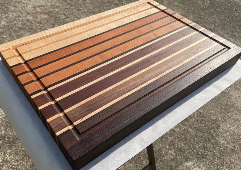 May include: A rectangular wooden cutting board with a striped design. The board features alternating stripes of light and dark wood tones, including shades of brown, tan, and black. A recessed groove runs along the edge of the board.