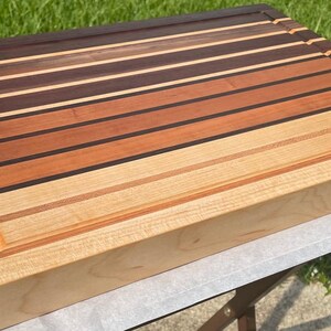 May include: A rectangular wooden cutting board with a variety of wood tones, including dark brown, reddish-brown, and light tan. The board has a juice groove around the edge and is made of multiple wood strips.