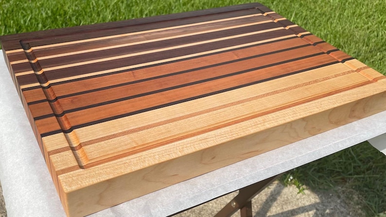 May include: A rectangular wooden cutting board with a variety of wood tones, including dark brown, reddish-brown, and light tan. The board has a juice groove around the edge and is made of multiple wood strips.