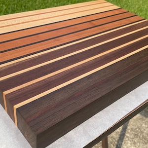 May include: A rectangular wooden cutting board with alternating stripes of light, medium, and dark brown wood. The board is made of various wood types, creating a visually appealing pattern. The board is sitting on a white surface.