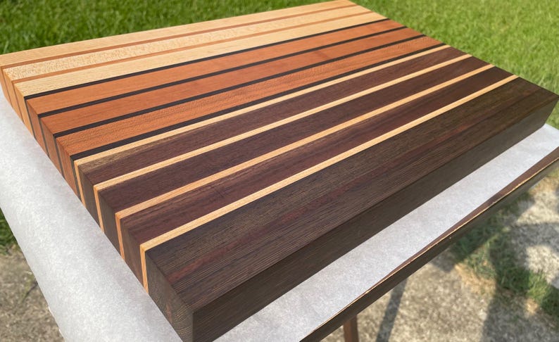 May include: A rectangular wooden cutting board with alternating stripes of light, medium, and dark brown wood. The board is made of various wood types, creating a visually appealing pattern. The board is sitting on a white surface.