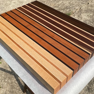 May include: A rectangular wooden cutting board with a striped design. The board features alternating strips of light, medium, and dark brown wood, creating a visually appealing pattern. The board is approximately 18 inches long and 12 inches wide.
