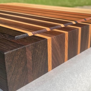 May include: A handcrafted wooden cutting board with a striped design. The board features alternating strips of dark brown, light brown, and tan wood. The end grain construction is visible, and the board has a rectangular shape.