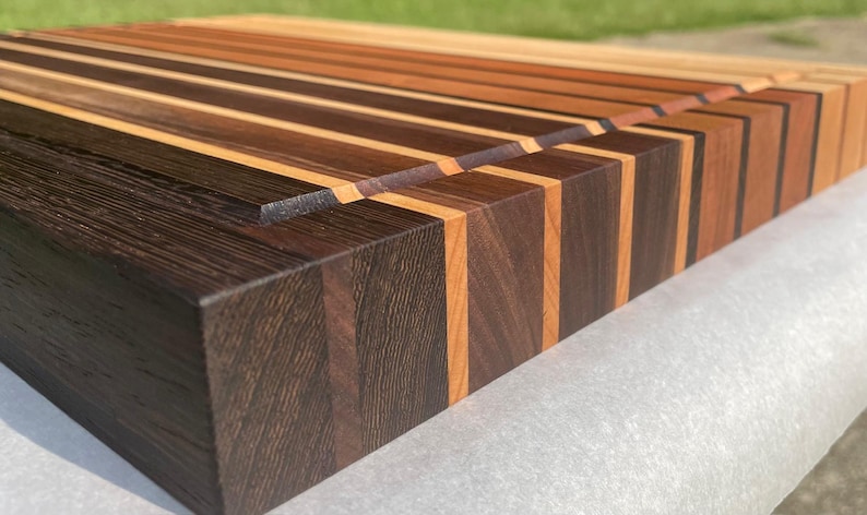 May include: A handcrafted wooden cutting board with a striped design. The board features alternating strips of dark brown, light brown, and tan wood. The end grain construction is visible, and the board has a rectangular shape.