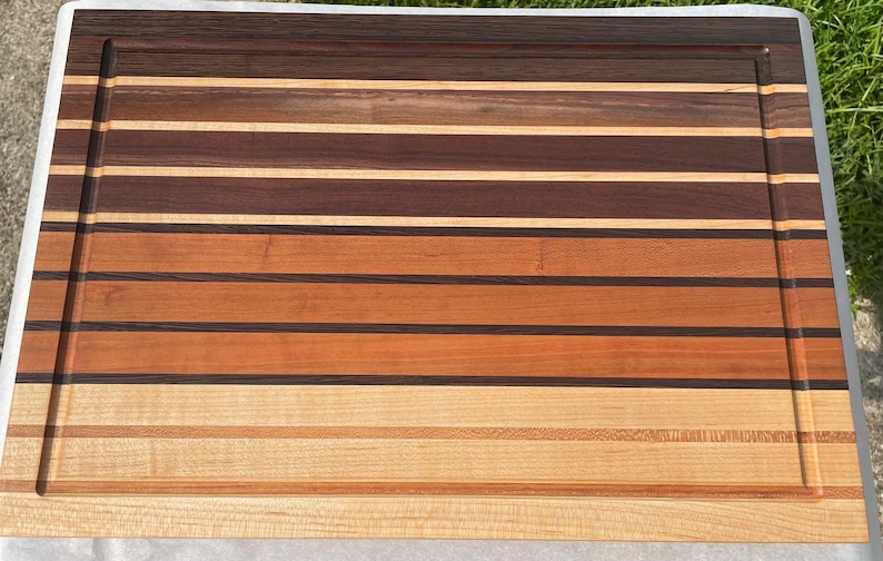 May include: A rectangular wooden cutting board with a variety of wood tones, including light tan, medium brown, and dark brown. The board has a juice groove around the perimeter and is made of multiple horizontal strips of wood.