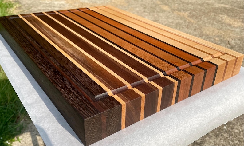 May include: A rectangular wooden cutting board with a striped pattern. The board features alternating strips of light and dark wood, creating a visually appealing design. A groove runs along one side. The board is likely used for food preparation.