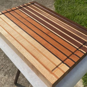 May include: A rectangular wooden cutting board with a variety of wood tones, including light, medium, and dark browns. The board has a juice groove and is constructed with alternating strips of wood. The board is approximately 18 inches long and 12 inches wide.