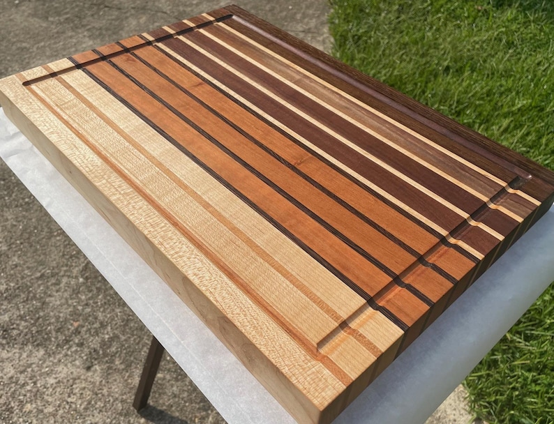 May include: A rectangular wooden cutting board with a variety of wood tones, including light, medium, and dark browns. The board has a juice groove and is constructed with alternating strips of wood. The board is approximately 18 inches long and 12 inches wide.