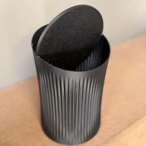 May include: A black cylindrical container with a textured lid, partially open. The container has vertical ridged detailing. The lid is round and has a rough, matte finish. The container is sitting on a wooden surface.
